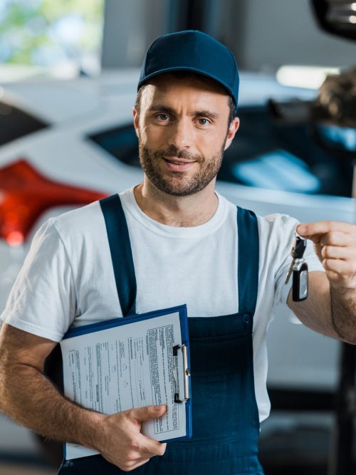 happy car mechanic looking at camera and holding clipboard and key near car e1653233732554 Cheapest Towing Near Me Corona, California